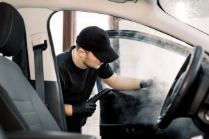 Auto cleaning service and detailing concept. Handsome Caucasian man in black uniform cleaning interior of the car with hot steam cleaner. Selective focus